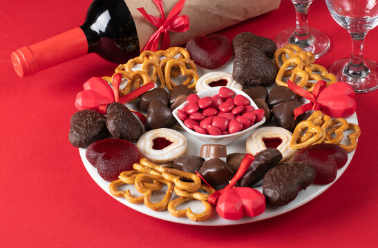 Plate of assorted heart-shaped sweets, two glasses and bottle wine for Valentine's Day on red surface, Close up