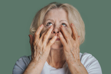 Senior woman applyin nourishing cream under her eyes on green studio background. Domestic beauty...