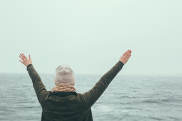 Freedom: back view of the happy woman standing on the shore of the sea in cold day and raising hands up.