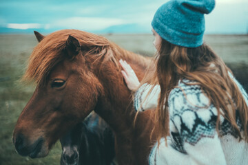 Back view of woman in lopapeysa on the nature. Tourist female pats the beautiful brown horse in the mountains in Iceland. - obrazy, fototapety, plakaty