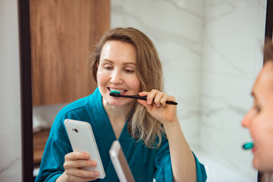 Social media affect. Young happy woman brushing teeth and looking at phone screen in stylish bathroom at mirror