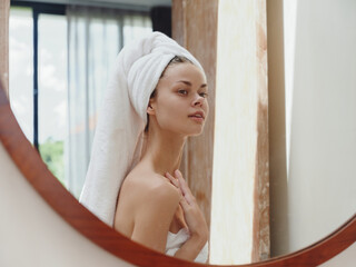 A beauty woman stands in front of a mirror after a shower in a towel on her head looks at her reflection and makes a facial massage against aging, beauty facial skin care smile, anti acne
