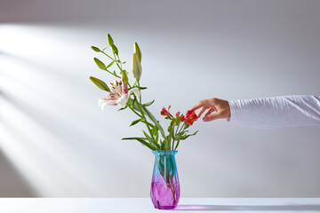 a man's hand touches a bouquet of flowers in a colored glass vase standing on a white table against the background of a white wall with shadows from the sun's rays