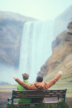 Back view of young couple sitting on the bench near the Skogafoss waterfall in Iceland. Man raised hands up near woman.