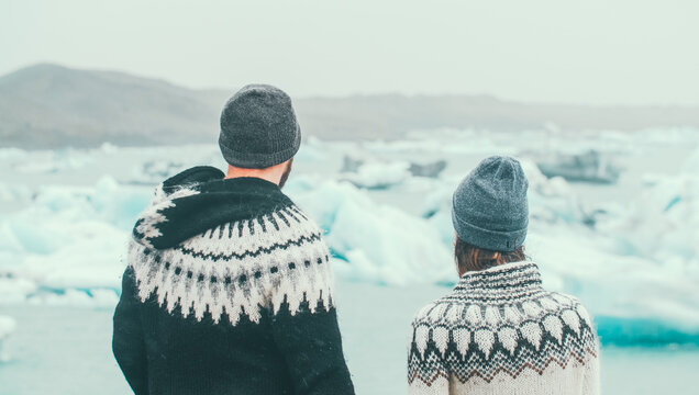 Back view of young tourists couple in lopapeysa sweaters standing in ice lagoon and looking at the glaciers in Iceland.