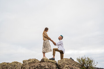 A young couple stands on a rocky cliff by the ocean, with the man down on one knee proposing to the...