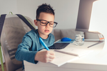 Little boy sitting in his room at a desk next to a computer and doing homework preparing for school.