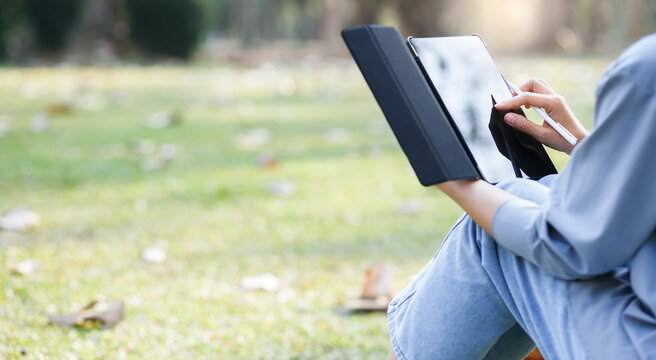 Young asian woman with digital tablet sits on the grass in the park on a sunny day. she using tablet online working, remote work and communication in social networks and working part time