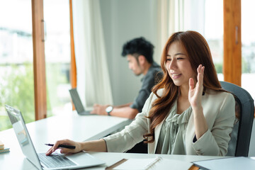 Beautiful Asian businesswoman is on video call with customer on laptop in office and has a negotiating gesture smiling and happy face.