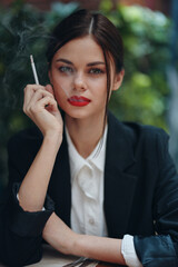 Obraz premium Stylish fashion woman portrait sitting in a cafe at a table and smoking a cigarette releasing smoke from her mouth with red lipstick, a bad habit, a smile with teeth and a thoughtful look