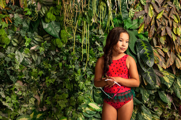 Asian little girl in red swimsuit with sunglasses in hand at tropical greenery, looking away. Suntan small lady kid model in swimwear. Childhood vacation concept. Copy text space for advertising