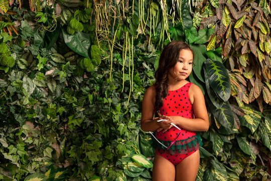 Asian little girl in red swimsuit with sunglasses in hand at tropical greenery, looking away. Suntan small lady kid model in swimwear. Childhood vacation concept. Copy text space for advertising