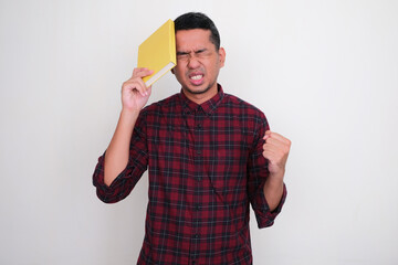 Adult Asian man showing stress expression while holding a book