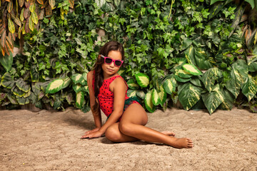 Asian little girl in red swimsuit in sunglasses sitting on sandy beach at tropical greenery,...