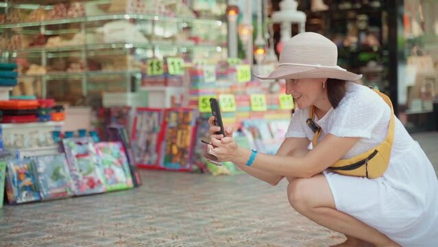 A woman in a white dress, squatting along the shopping malls of a tourist city, photographs an unknown object using a camera and an application on a modern mobile phone. Women's Wanderlust