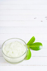 cosmetic cream in glass pot and leaves on white wood
