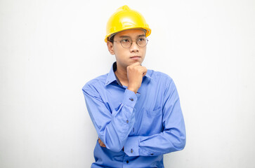 Asian construction worker in blue shirt and yellow safety helmet is seriously thinking with a wondering and thinking gesture and expression.