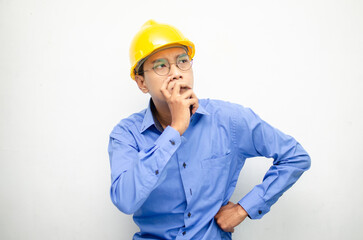 Asian construction worker in blue shirt and yellow safety helmet is seriously thinking with a wondering and thinking gesture and expression.