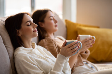 Satisfied european millennial ladies with closed eyes in sweaters with cups of coffee sit on sofa...