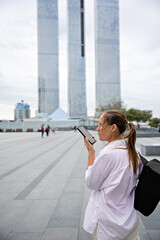 A girl with glasses stands with her back and looks at a big city, holding a phone in her hands