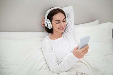 Relaxed young woman sitting in bed with phone and headphones
