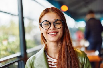 Woman hipster with red hair and glasses sits in town in cafe smile with teeth looking at camera, woman freelance blogger close-up