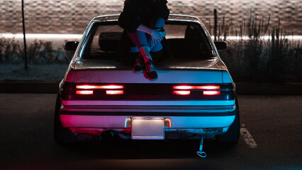 young woman is sitting on the trunk of a drift car . neon light. Night photo - obrazy, fototapety, plakaty