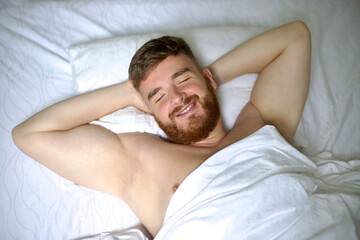 Fototapeta na wymiar Happy young handsome man is waking up in the morning in bed in bedroom, stretching his arms, body, smiling.