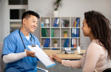 Happy middle aged korean man doctor giving signing contract to black millennial woman patient in...