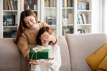 Cheerful european millennial lady closes eyes to her friend and gives gift box in living room...
