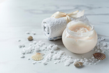 Fototapeta na wymiar Open jar of skin cream, towel and scattered sea salt and shells on the light marble background. Spa composition. Copy space
