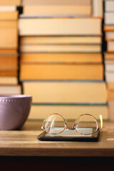 Cup of tea or coffee, e-reader and reading glasses on the table. Pile of books in the background. Selective focus.