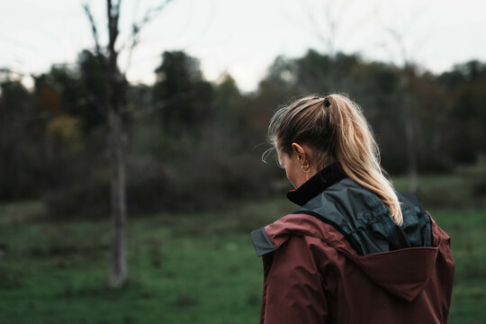 Blond girl walks in an empty meadow. Backview of young hiker strolling alone in countryside. Cloudy day, melancholy and loneliness. - Powered by Adobe
