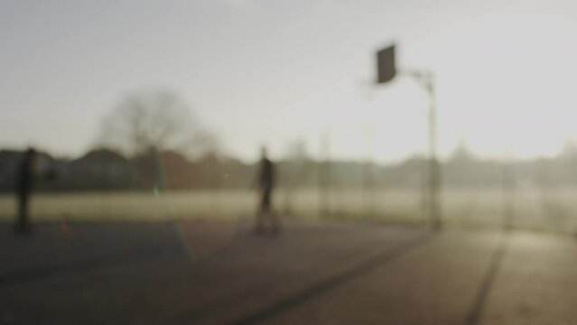 Panning shot of blurred background of two silhouette people shooting a basketball on an outdoor court, in slow motion