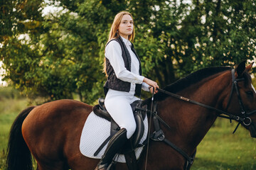 Young beautiful blonde woman jockey rides a brown horse in a meadow at sunset in summer. Preparing...