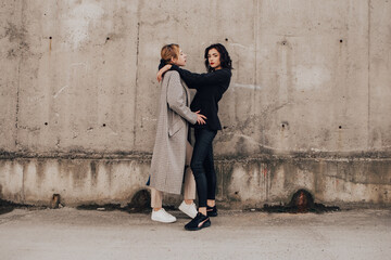 Two pretty women friends posing near gray wall. Couple of gay lesbian girls hugging embracing together girlfriends, dressed casual outfits, have a date. LGBT concept. Fashion, make up, hairstyle
