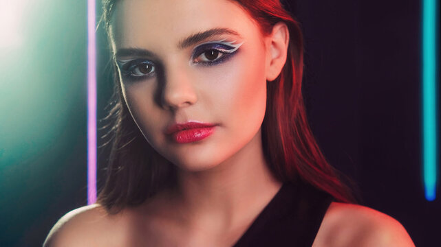90s portrait. Retro look. 2000s fashion model. Blue color neon light closeup of teen girl face with purple eyeshadow pink lips artistic makeup on dark black.