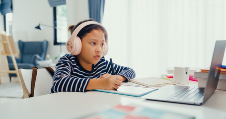 Asian toddler girl with sweater wear headphone sit front of desk with notepad use magic pen focus on writing do homework, finger counting number math online learning course from laptop at home.