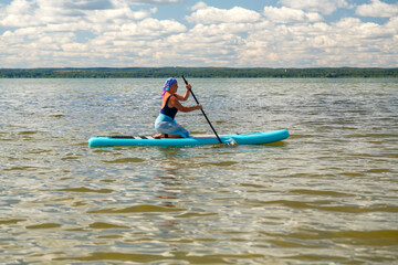A woman in a scarf on her head and in a pareo on a SUP board with an oar swims in the lake on a sunny day.