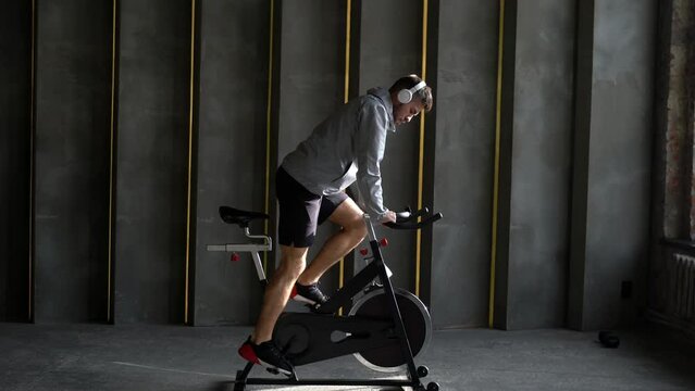 Pumped up male athlete listening audio book during intensive cardio workout for training leg muscles while cycling on exercise bike, determined man in sportswear keeping goals while riding in gym
