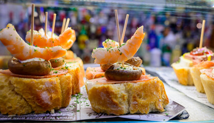 Fototapeta premium White board with typical snack of Basque Country, pinchos or pinxtos skewers with small pieces of bread, courgette, sea food, eggs, cheese, served in bar in San-Sebastian or Bilbao