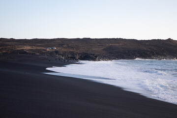 Wide shot of a black sand beack with the mountains at the background and the white waves arriving to the edge