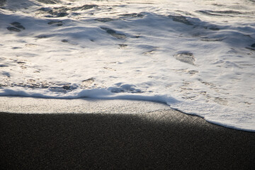 Close up of a white wave at the edge of a black sand beach during the golden hour 