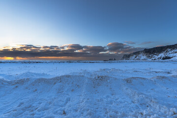 Naklejka na ściany i meble Landscape of the beach area Totally snow covered black sand beach with golden sunrise light under the clouds