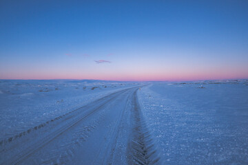 Road and flat snowy landscape with tire tracks going to the horizon with purple magical icelandic sunrise.