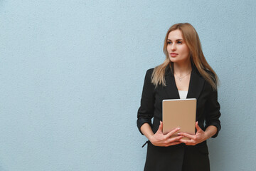 Blonde young business woman wearing black suit over blue plain background, carrying laptop