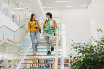 Naklejka na ściany i meble two women friends of colleagues walk up glass steps in coworking office. Female students discuss work project, creative work. marketing and finance. career and success, confident open people