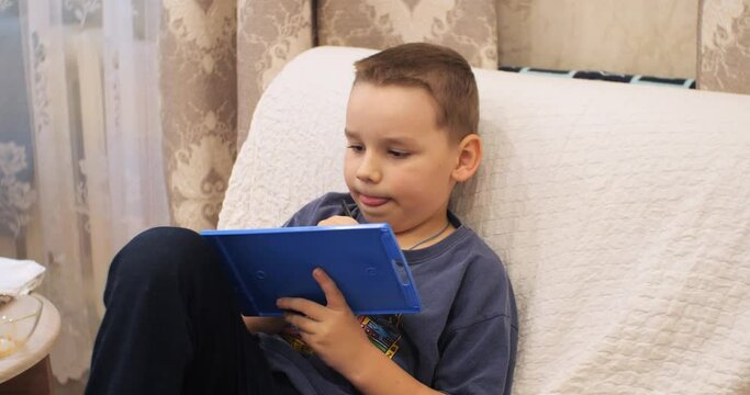 The child sits in a chair and enthusiastically draws on the tablet with a pen. A child with sincere emotions draws on a tablet on his knees while sitting in an armchair.