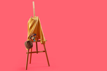 Easel with yellow sweater, beret and paint palette on red background