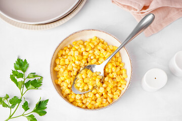 Bowl with canned corn, spoon and parsley on white table
